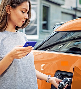 A woman holding a smartphone while she plugs in the charger to her electric car